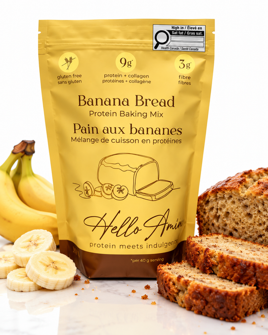 A stand-up pouch of Hello Amin Banana Bread Protein Baking Mix stands on a white marble surface with bananas, banana slices, and sliced banana bread around it. The pouch is golden yellow with black and brown text, and a Hello Amin brand logo is clearly visible.