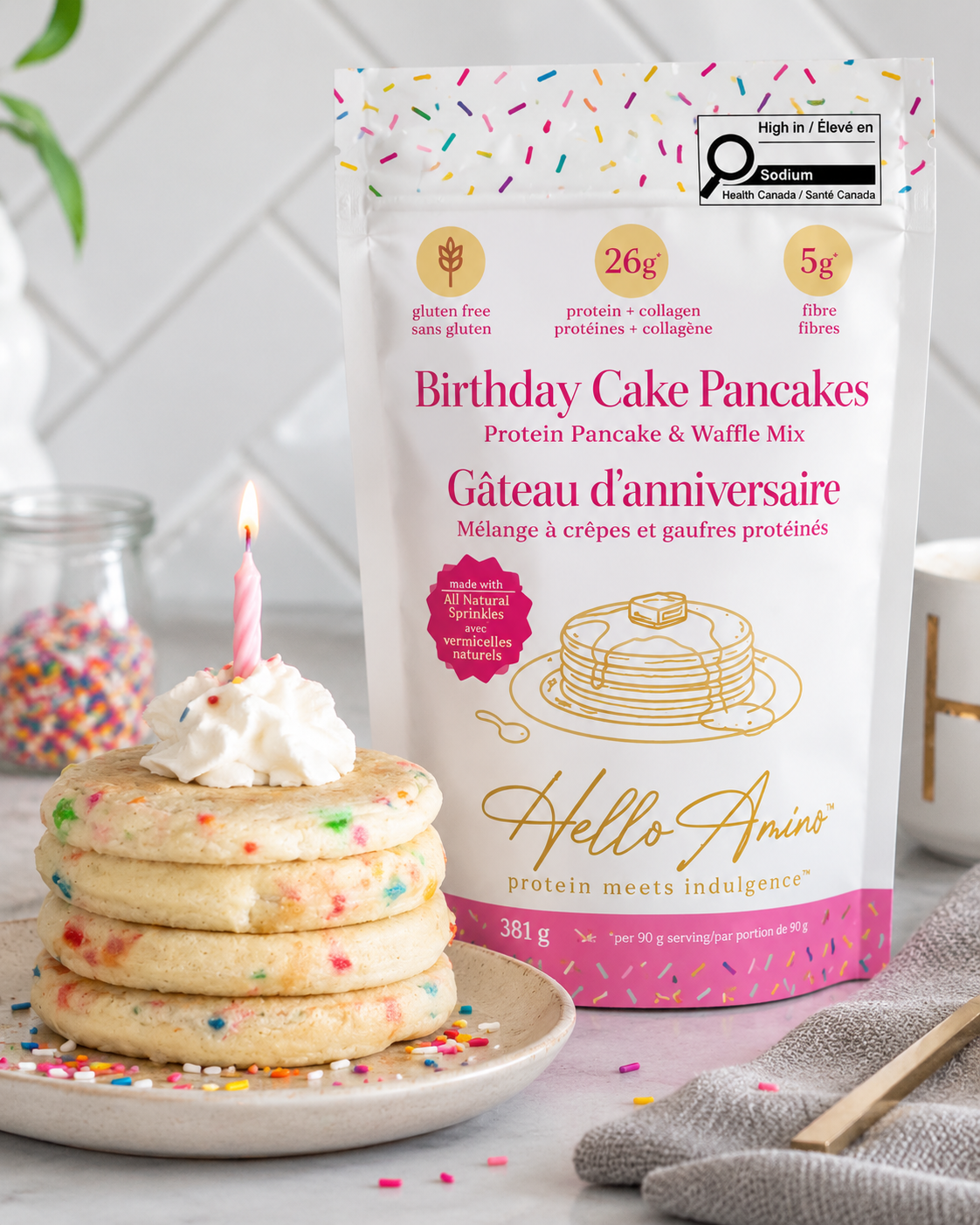 A bag of HelloAmino's Birthday Cake Pancakes protein pancake and waffle mix stands next to a stack of four birthday pancakes topped with whipped cream and a single lit candle on a plate, with colorful sprinkles scattered around in a bright kitchen setting.