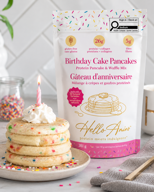 A bag of HelloAmino's Birthday Cake Pancakes protein pancake and waffle mix stands next to a stack of four birthday pancakes topped with whipped cream and a single lit candle on a plate, with colorful sprinkles scattered around in a bright kitchen setting.
