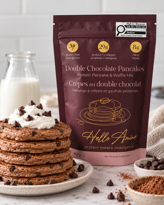 A stand-up pouch of HelloAmino's Double Chocolate Pancakes protein pancake and waffle mix stands on a marble surface in front of a stack of chocolate chip pancakes topped with whipped cream. A glass bottle of milk, cocoa powder, and chocolate chips are arranged around it in a bright kitchen-like setting.