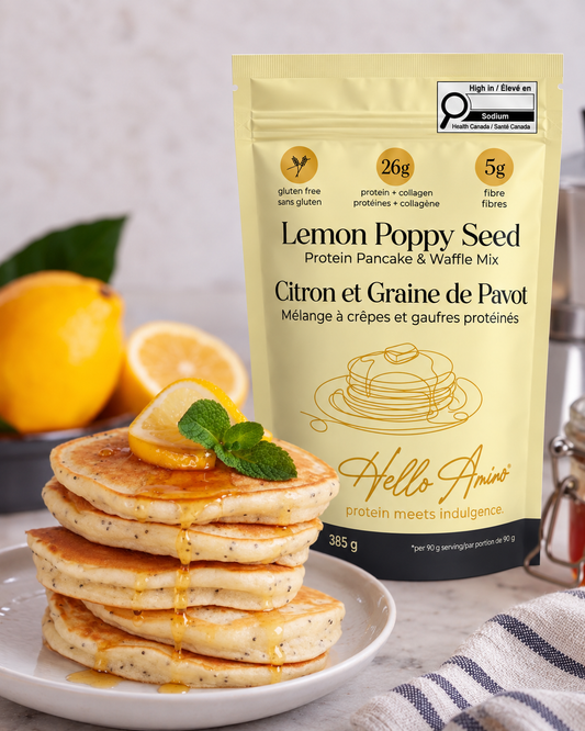 A stand-up pouch of HelloAmino Lemon Poppy Seed Protein Pancake & Waffle Mix stands on a countertop beside a plate of stacked pancakes topped with syrup, lemon slices, and mint, with whole lemons and a cloth in the background.