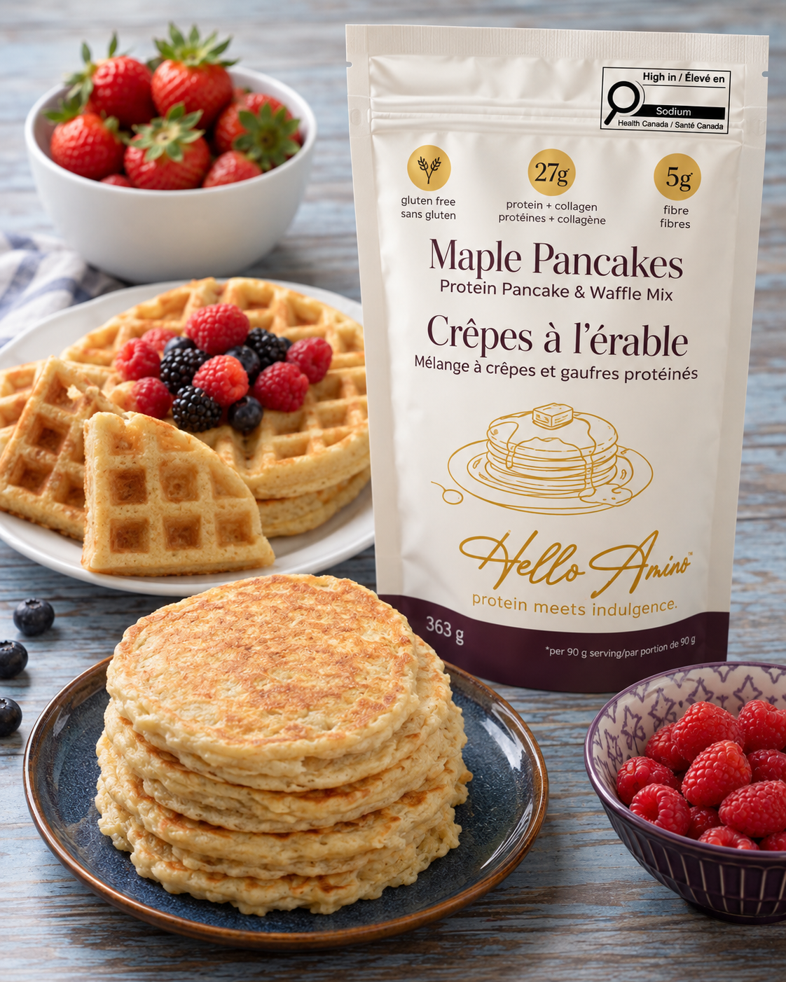 A bag of HelloAmino Maple Pancakes Protein Pancake & Waffle Mix stands on a rustic blue wooden table, surrounded by plates of golden-brown pancakes, fresh strawberries, and berries. The packaging features a white background with gold and dark purple text and graphics.