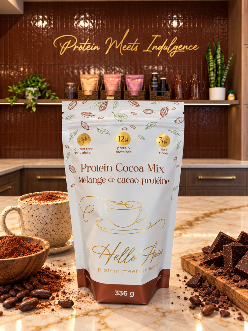 A stand-up pouch of HelloAmino Protein Cocoa Mix stands on a marble countertop in a their Calgary Sip Bar location, surrounded by cocoa powder, cocoa beans, chocolate chunks, and a cup of hot cocoa. The background features a dark wood tiled wall with gold script reading 'Protein Meets Indulgence'.