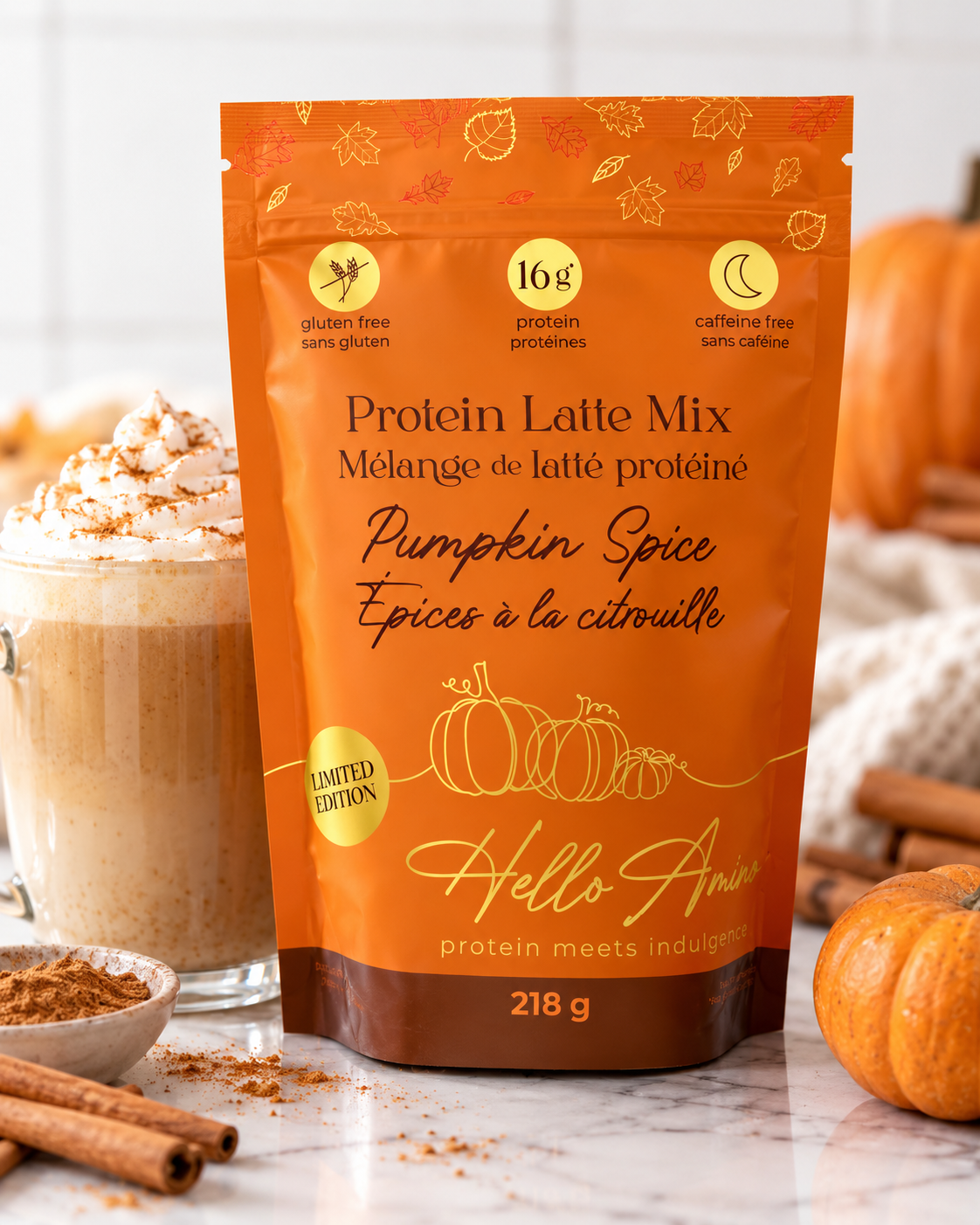 A stand-up pouch of HelloAmino Pumpkin Spice Protein Latte Mix in an orange package, styled on a marble surface with a pumpkin, cinnamon sticks, and a protein latte in the background.