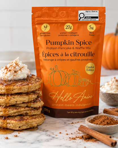 A bag of HelloAmino Pumpkin Spice Protein Pancake & Waffle Mix stands on a light surface beside a stack of pumpkin-spice pancakes topped with whipped cream and drizzle, with cinnamon sticks and a small bowl in the background.