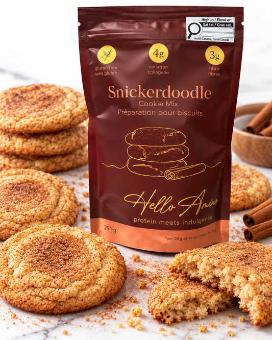 A stand-up pouch of HelloAmino Snickerdoodle Cookie Mix stands on a marble surface surrounded by cinnamon-sugar-coated cookies, with a whole cookie and a broken piece in front. The package is dark brown with gold and white text and graphics, and the scene is styled with cinnamon sticks and cookies.