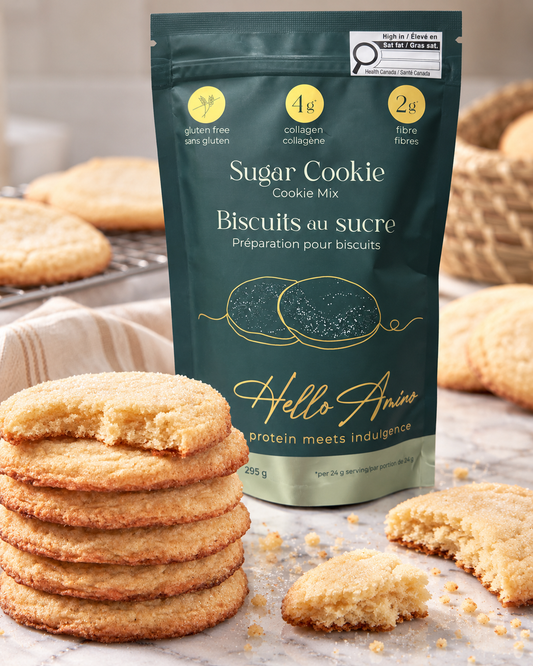 A stand-up pouch of HelloAmino Sugar Cookie Mix stands on a marble surface with baked cookies around it. The pouch is dark teal with yellow and white text and graphics, and the background shows golden-brown cookies and crumbs.